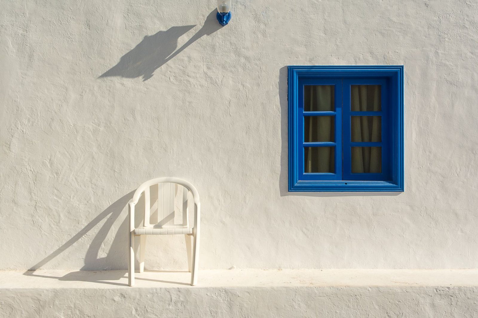 A sunlit white Mediterranean wall with a blue window and a simple chair